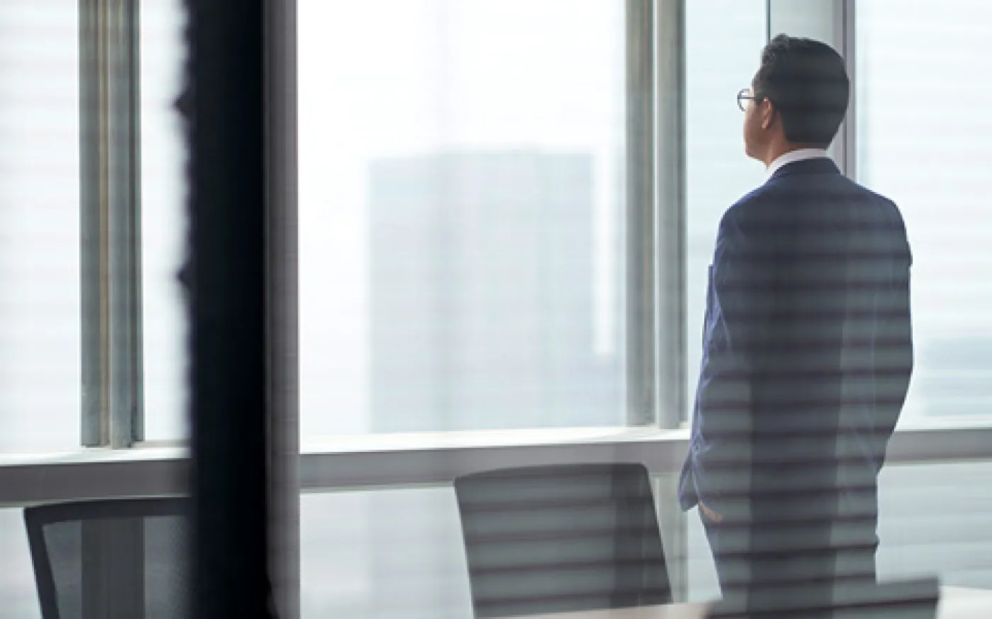 Business professional in a suit standing by office window, looking at city skyline in a modern workspace.